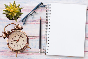 Retro alarm clock,blank notebook,glasses and flower on a wooden table. Photo in retro color image...