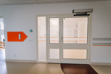 A hospital corridor with an orange directional arrow sign, frosted glass doors, and a clean, modern...