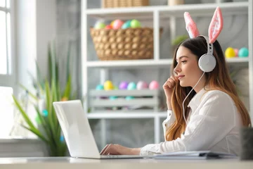 Foto auf Alu-Dibond Side view portrait of businesswoman sitting at the home office desk and looking at laptop screen. She is wearing bunny ears headphones. Colorful easter eggs in the background. © zphoto83