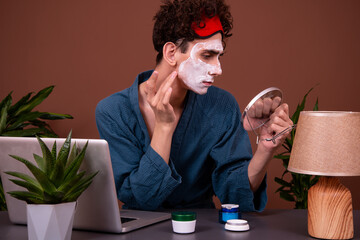 Funny man with cream on his face. Spa treatments. Skin care. Morning procedures before starting work. Brown background. The guy is looking at the camera.