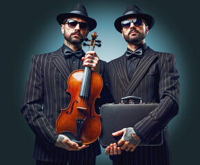 Tattoed mafia brothers posing for a picture while holding a violin and a bag in their hands.