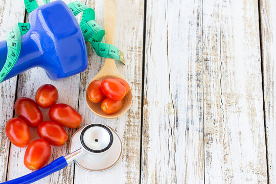 Blue stethoscope and dumbbell with red tomatos on white wooden table