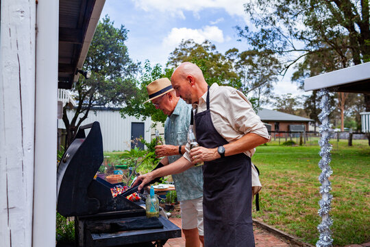 men cooking barbeque lunch in backyard together holding beers in hand