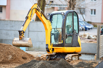 Tracked orange mini excavator digger near road with green trees and concrete fence on background with copyspace.