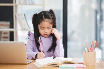 A young girl is sitting at a desk with a laptop and a book