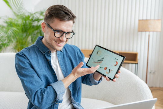 Smiling man in glasses presenting tablet with graphs, in a light, stylish room.