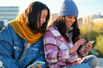 Two multiracial female friends laughing and sharing something on the smartphone at street in autumn.