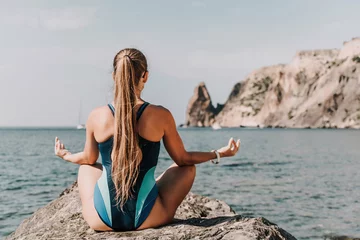 Gordijnen Yoga on the beach. A happy woman meditating in a yoga pose on the beach, surrounded by the ocean and rock mountains, promoting a healthy lifestyle outdoors in nature, and inspiring fitness concept. © svetograph