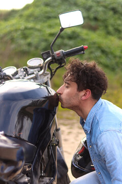 Curly haired Caucasian young man alone, kissing motorbike with affection, side view in close-up. Concept of love for the motorcycle, vertical image