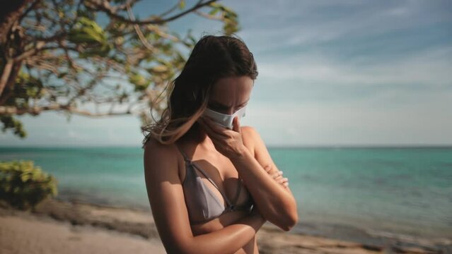 Woman with COVID-19 medical mask stands alone on a sunny beach. Girl in mask got sick coughs while have holiday vacation on tropical island. Coronavirus prevention concept. Covid pandemic. Slow motion