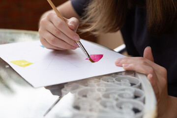 Cut-out of a teenage girl's hands painting with watercolors with a brush on a white sheet at...