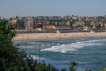 Fototapeta na wymiar Iconic Bondi Beach and Bondi Pavilion, in Sydney, Australia. People in the sand, water and carrying surf boards. Green shrubs in the foreground.