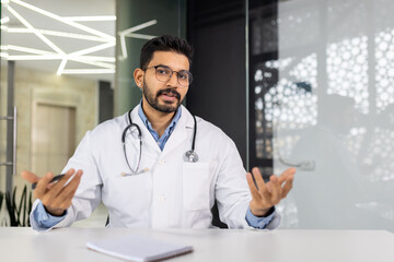 A doctor is sitting at a desk with a clipboard and a pen. He is wearing a white lab coat and glasses. The scene appears to be a medical office or examination room