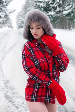 a sexy beautiful girl in a red checkered shirt, a hat with earflaps, knitted red mittens with bare legs stands looking down against the backdrop of a snow-covered forest road
