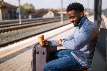 Happy man with suitcase and  coffee looking a clock while sitting on a bench at the railway station. - 765634964