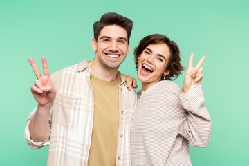 Cheerful, beautiful couple hugging, showing victory gestures with fingers, isolated on turquoise - obrazy, fototapety, plakaty