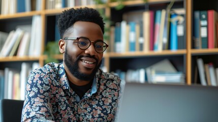 A young man is sitting in a library, smiling while looking at his laptop. He is wearing glasses and has a beard.