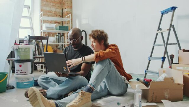 Young diverse couple sitting on the floor in the living room under renovation, looking for interior design ideas on the Internet and having discussion. Zoom Shot