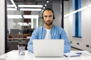 Fototapeta na wymiar Portrait of a serious young hispanic male specialist wearing a headset, sitting at a desk in a busy office, working on a laptop, looking intently at the camera