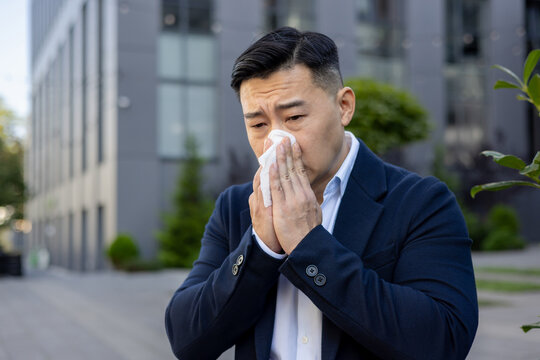 Close-up photo of a sick young Asian male businessman sitting on a bench near an office building and wiping his nose with a napkin from a runny nose