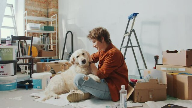Young joyous woman petting cute golden retriever dog in living room with ladder, boxes and paint buckets while doing home renovation. Zoom Shot