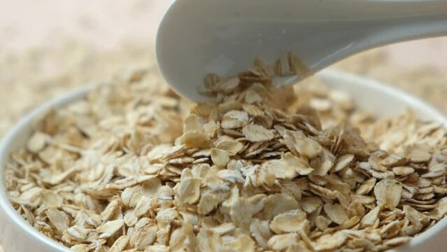 Uncooked dry oat flakes pouring from spoon into plate slow motion close-up.