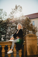A woman is sitting on a stone railing with her legs crossed. She is wearing a black coat and a white scarf. The scene is set in a city with a building in the background. The woman is enjoying the view