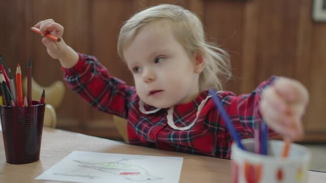 A little charming girl sitting at the desk chooses a colored pencil for drawing. Baby girl learning colors and doing art.