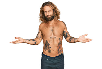 Handsome man with beard and long hair standing shirtless showing tattoos clueless and confused with open arms, no idea concept.
