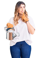 Young caucasian woman wearing gloves holding watering can serious face thinking about question with...