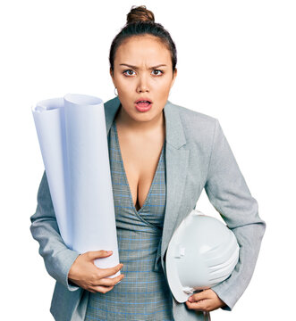 Young hispanic girl holding safety helmet and blueprints afraid and shocked with surprise and amazed expression, fear and excited face.