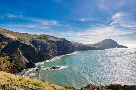The photo depicts a cliff overlooking a bay and mountains in Madeira. The sky is blue, and the water is blue. In the foreground, there are trees and bushes. A person stands on the cliff.