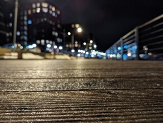 A wooden walkway illuminated by automotive lighting leading to a city at night