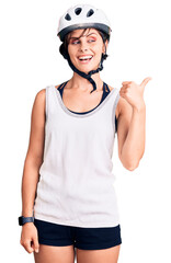 Beautiful young woman with short hair wearing bike helmet smiling with happy face looking and pointing to the side with thumb up.