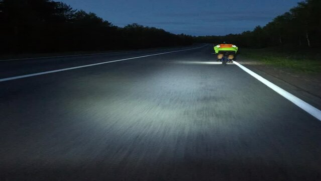 A person is riding a bike down a highway at night with automotive lighting illuminating the road surface, asphalt, and landscape. The sky is dark and the cars exterior reflects in the windshield