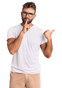 Young hispanic man wearing casual clothes and glasses asking to be quiet with finger on lips pointing with hand to the side. silence and secret concept.