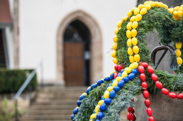 Osterbrunnen mit gelben, roten und blauen Ostereiern aus Kunststoff vor historischem Gebäude