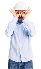 Young handsome man wearing architect hardhat doing ok gesture like binoculars sticking tongue out, eyes looking through fingers. crazy expression.