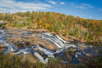 In a backdrop of fall foliage and blue sky, the scenic Towaliga River cascades downstream over granite bedrock. Captured from Tranquility Trail in High Falls State Park in Georgia. © Donna Bollenbach