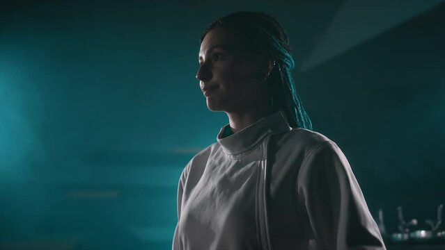 Side tilt lowkey footage of female fencer zipping up her vest in dark smokey room before training