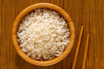 Cooked white rice in a dark wood bowl next to chopsticks isolated on bamboo matt from above.
