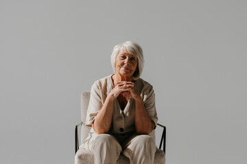 Relaxed gray hair senior woman sitting in comfortable chair and looking at camera in sunny studio