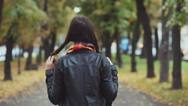 Young woman walking down tree-lined city street on autumn day, playfully twirling her hair