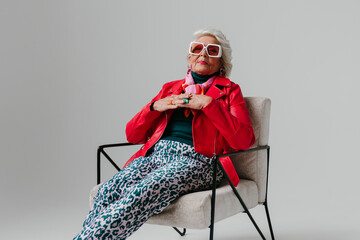 Cool elderly woman in fashionable clothing sitting in comfortable chair on grey background