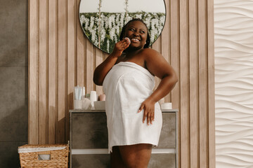Happy plus size African woman covered in towel enjoying beauty treatment at the domestic bathroom - 765788563