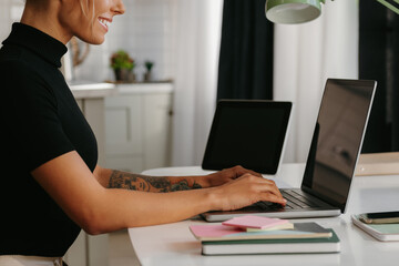 Side view of smiling young woman sitting at the desk and using laptop while working at home