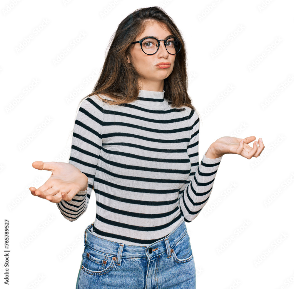 Sticker Young beautiful teen girl wearing casual clothes and glasses clueless and confused expression with arms and hands raised. doubt concept.