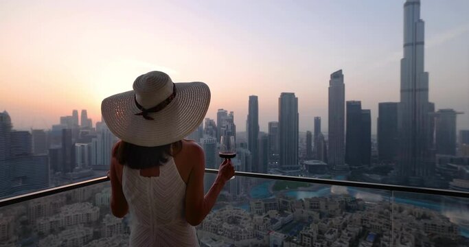 A elegant luxury woman enjoys the beautiful sunset view behind the modern skyline of Downtown Dubai, UAE, with a drink