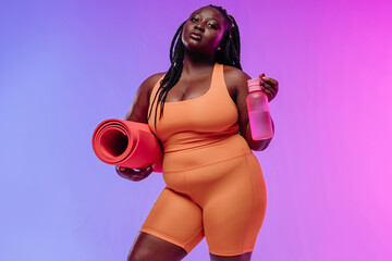 Active young voluptuous woman in sportswear carrying exercise mat and smiling on pink background