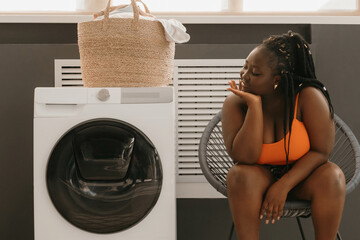 Bored curvy African woman waiting for clean clothes while sitting in chair near the washing machine in laundry - 765791518
